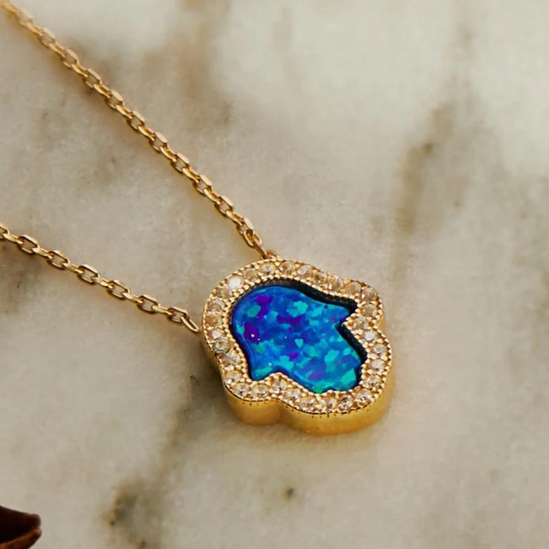 A close up of the Hamsa Opalite Turquoise Blue Gold Necklace against a marble background.